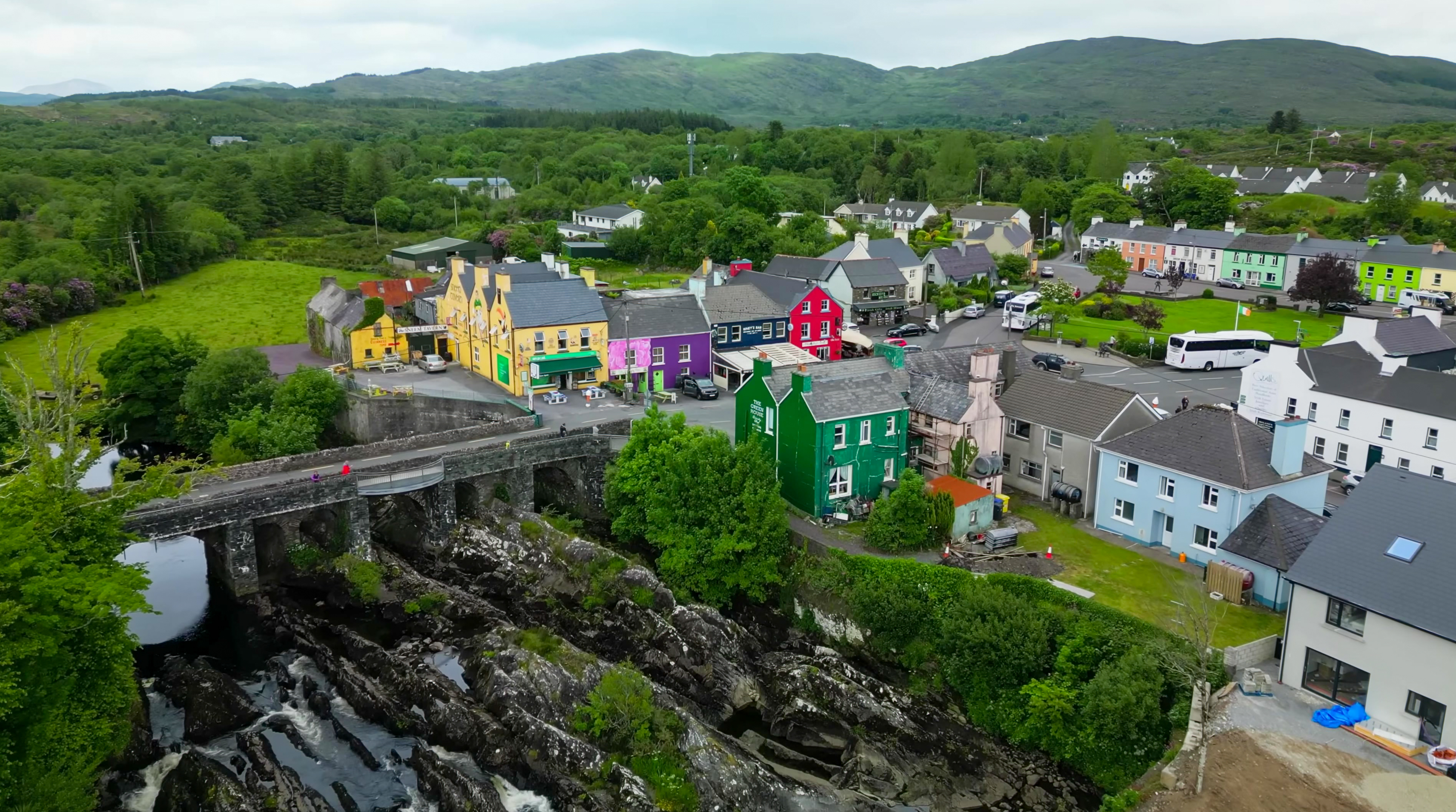 Aerial drone view of Sneem village in County Kerry, Ireland, revealing a cluster of vividly painted shopfronts and houses in yellow, purple, red, and green alongside a historic multi-arch stone bridge spanning a rocky river cascade. The compact village centre is surrounded by lush green hills, dense woodland, and rolling mountains under a soft overcast summer sky. The scene captures the charm and colour of a traditional Irish village on the Iveragh Peninsula.